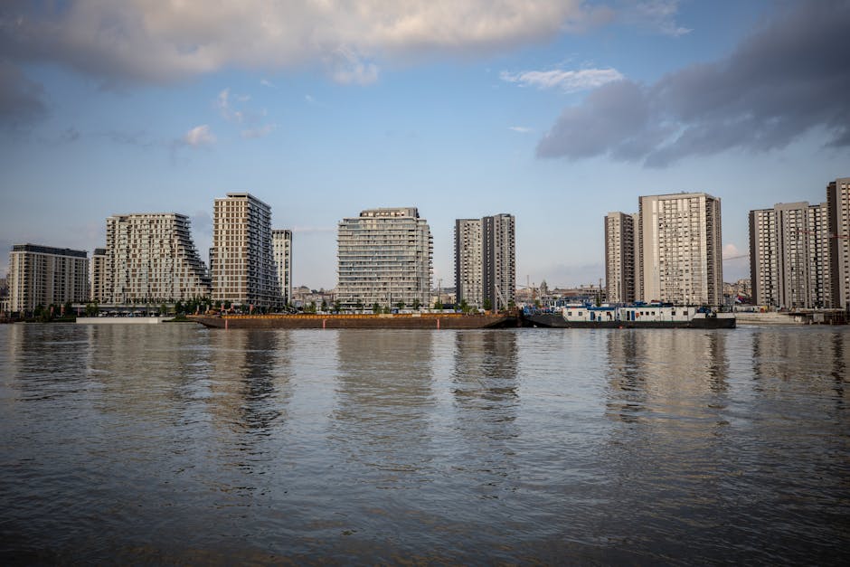 A modern, multi-storey residential building with a yellow brick façade and numerous glass-fronted balconies, located along a waterfront with boats docked nearby. The building features large circular 