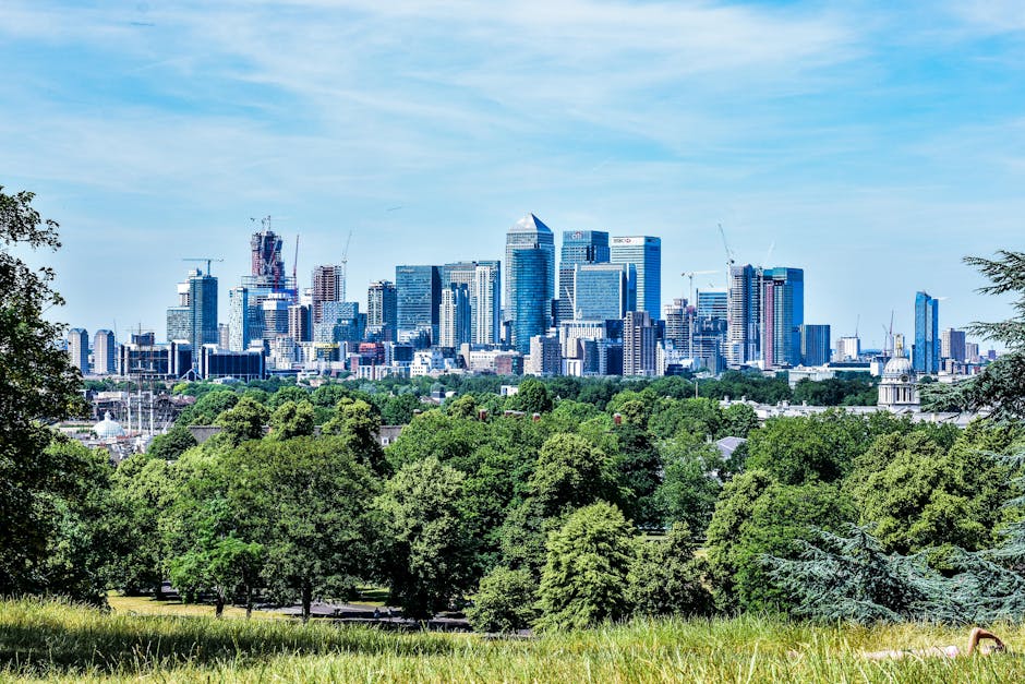 Photograph of a modern urban skyline in Canary Wharf featuring several high-rise office buildings with glass facades reflecting the sky, alongside a red train crossing a bridge over a calm river. The 