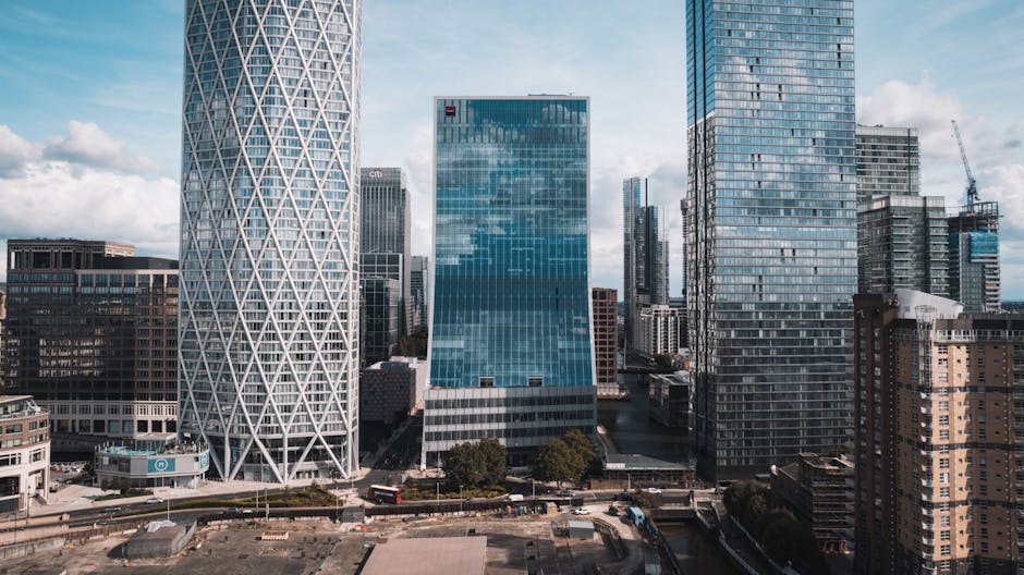 A monochrome image of the Canary Wharf skyline featuring tall modern skyscrapers with glass facades reflecting clouds in the sky. The buildings vary in height and architectural style, including one wi